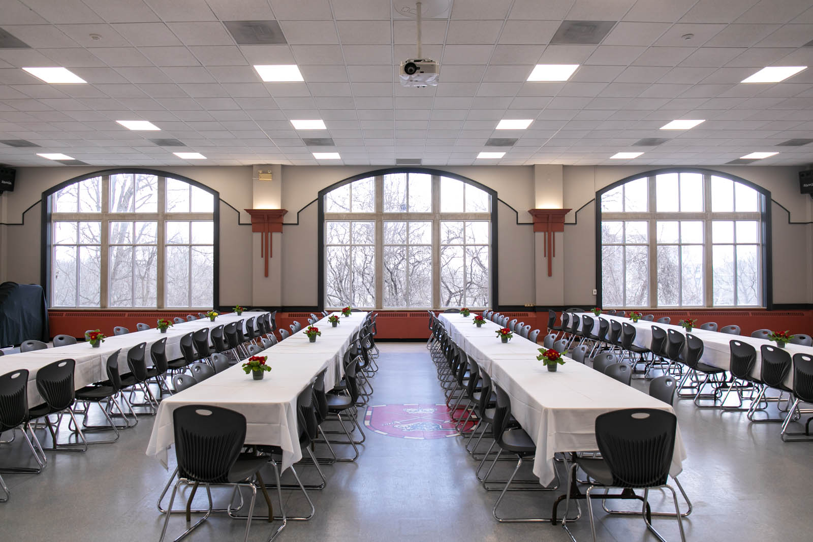 A large room with three bay windows at the back set with four long rows of tables and banquet seating for 120 people