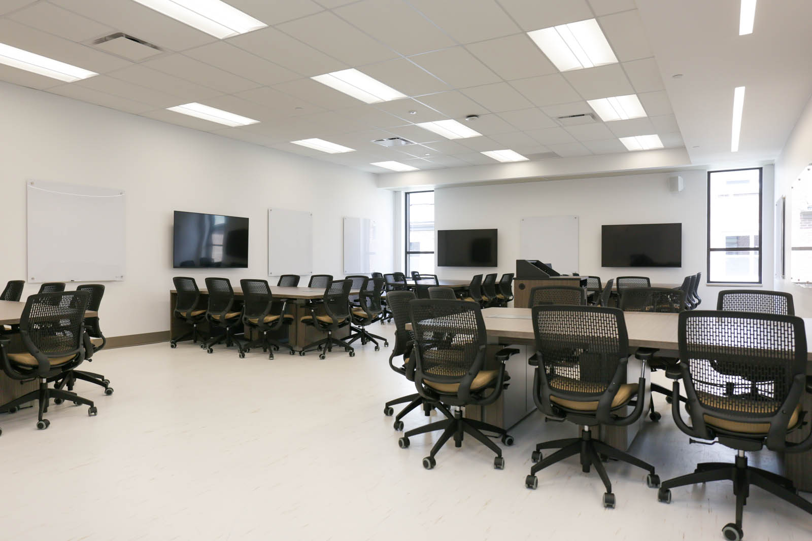 A bright classroom with several TVs and whiteboards as well as a podium and 5 tables with seating for 40 people